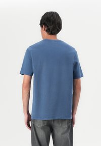 Camiseta de algodón azul de manga corta con cuello redondo, corte relajado y detalles de costura sutiles. Se lleva con jeans grises.