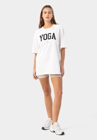 Camiseta blanca de gran tamaño con "YOGA" en letras negras en negrita, combinada con pantalones cortos beige y zapatillas ligeras, de pie sobre un fondo neutro.