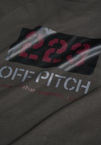 Czarna koszulka z ciemnoszarego materiału o gładkiej teksturze, z odważnym nadrukiem graficznym: czerwone cyfry "223" oraz tekst "OFF PITCH" w kolorze srebrnym.