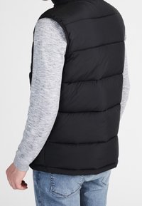 Svart vadderad väst med hög krage, med horisontell sömnad och en slät textur, bärs över en grå långärmad skjorta och blå jeans.