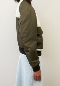 Olijfgroene bomberjack met een witte nepbontkraag, geribbelde manchetten en een zijzak; gladde textuur en crop-top ontwerp.