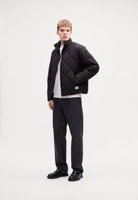 Young man wearing black quilted jacket, white shirt, dark jeans, and black shoes standing against plain white background.
