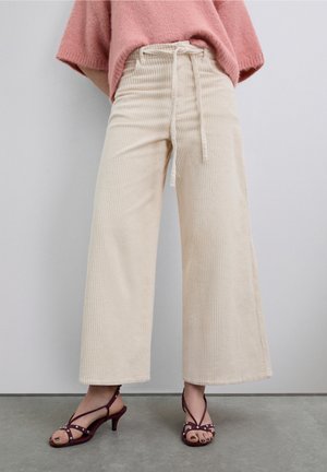Pantalon large en velours côtelé beige avec taille à cordon, associé à des sandales ouvertes bordeaux avec des accents décoratifs.