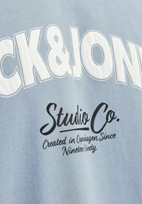 Camiseta de algodón azul claro con gran texto blanco "Jack & Jones" y texto negro "Studio Co." con acentos decorativos y una textura suave.
