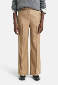 Persona che indossa pantaloni beige a gamba larga, scarpe nere e un maglione grigio con i polsini di una camicia bianca visibili, in piedi davanti a uno sfondo neutro.