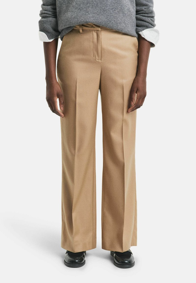 Persona che indossa pantaloni beige a gamba larga, scarpe nere e un maglione grigio con i polsini di una camicia bianca visibili, in piedi davanti a uno sfondo neutro.