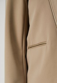 Beige blazer fabric with a smooth texture, featuring a single chest pocket and subtle piping detail along the pocket edge.
