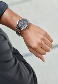 Montre en acier inoxydable argenté avec un cadran rond gris texturé, des fonctionnalités de chronographe, et un bracelet en maillons métalliques, portée sur un poignet.
