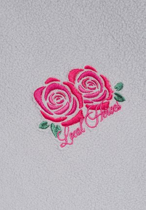 Polarowa tkanina w jasnoszarym tle z dwoma haftowanymi różowymi różami i zielonymi liśćmi, z napisem "Lokalni Bohaterowie".