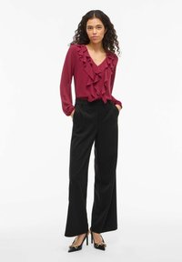 Bordowa bluzka z falbanami i dekoltem w serek, zestawiona z czarnymi spodniami o szerokich nogawkach. Modelka ma na sobie czarne szpilki z ozdobnymi akcentami.