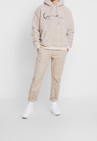 Beige Fleece-Hoodie mit einer Fronttasche und schwarzem Grafikttext. Kombiniert mit beigen Chinos und weißen Sneakers, mit aufgekrempelten Ärmeln.