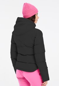 Schwarze Pufferjacke mit Kapuze, ausgestattet mit einem gesteppten Design und elastischen Bündchen. Kombiniert mit einer rosa Strickmütze und rosa Hosen.