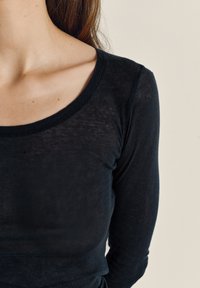 Žena koja nosi usku, prozirnu crnu majicu s dugim rukavima, na kojoj su vidljiva ključna kost i dio vrata te ramena, na neutralnoj pozadini.