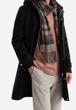 Muž nosící černý kabát s kapucí přes růžovou košili, béžové kalhoty a vícebarevnou kostkovanou šálu kolem krku. Ruce v kapse.