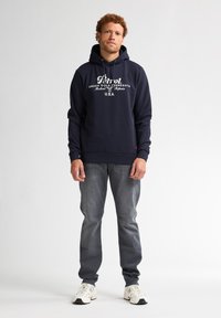 Námořnická modrá mikina s kapucí s předním grafickým textem, spárovaná s šedými džínami a bílými teniskami. Mikina má šnůrku a žebrované manžety.