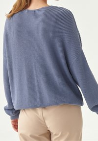 Femme portant un pull bleu ample et texturé ainsi qu'un pantalon beige, vue de dos sur un fond clair uni.