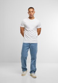 Weißes Baumwoll-T-Shirt mit rundem Halsausschnitt. Hellblaue Jeans mit seitlichen Druckknopfverschlüssen. Helle Sneakers mit beigen Akzenten. Neutraler Hintergrund.