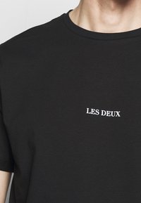 Černé bavlněné tričko s kulatým výstřihem, na hrudi má malé bílé textové logo "LES DEUX". Krátké rukávy, hladká textura.
