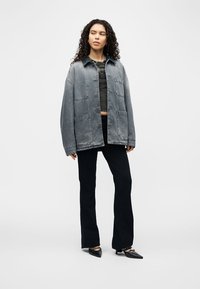 Femme aux cheveux bouclés portant une veste en denim grise oversize, un crop top noir, un pantalon noir et des talons noirs pointus, debout devant un fond blanc.