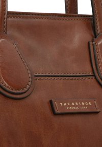 Borsa a mano in pelle marrone con una texture liscia, caratterizzata da due manici arrotondati e un'etichetta logo in rilievo dorato. I dettagli includono cuciture nere.