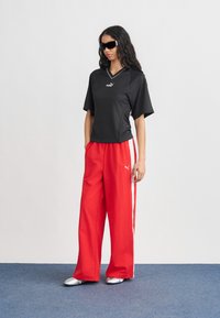 Γυναίκα με μαύρη μπλούζα Puma, κόκκινο παντελόνι φαρδιάς γραμμής με λευκές ρίγες στο πλάι, ασημένια παπούτσια και μαύρα γυαλιά ηλίου, στέκεται σε μπλε χαλί.