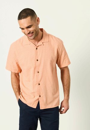 Uśmiechnięty mężczyzna z krótkimi włosami, ubrany w brzoskwiniową, wzorzystą koszulę na krótki rękaw zapinaną na guziki oraz ciemne spodnie, patrzy w dół z rękami w kieszeniach.