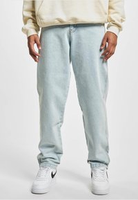 Blugi din denim albastru deschis, cu o croială relaxată, având cusături laterale și o glezna ușor conică, purtați cu adidași albi.