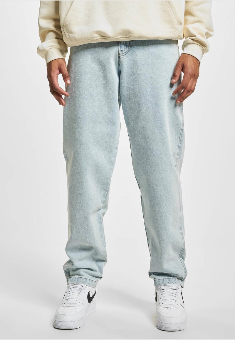 Blugi din denim albastru deschis, cu o croială relaxată, având cusături laterale și o glezna ușor conică, purtați cu adidași albi.