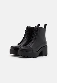 Botas de tobillo con plataforma de cuero negro, con cordones, tacones gruesos y tiradores, mostradas sobre un fondo blanco liso.
