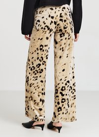 Weit geschnittene Hosen aus einem weichen, beigen Stoff mit einem schwarzen Fleckenmuster. Verfügen über zwei hintere Taschen und eine glatte Textur. Abgerundet mit schwarzen Schuhen mit Absatz.