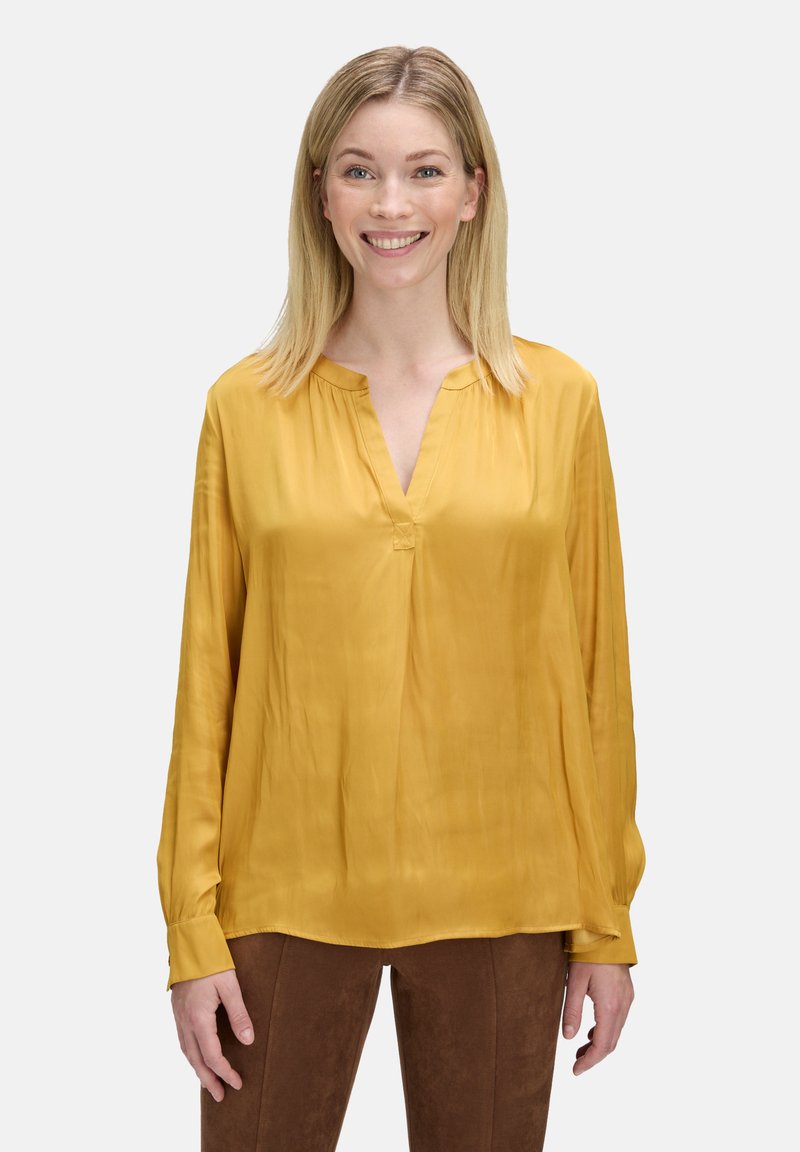 Lachende Frau mit glattem, blondem Haar, die eine senfgelbe Bluse und braune Hosen trägt und vor einem hellgrauen Hintergrund steht.