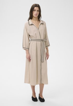 Mujer con vestido midi beige con ribete zigzag negro, botones en la parte delantera, cinturón con lazo, mangas tres cuartos y zapatos negros sin cordones sobre fondo blanco.