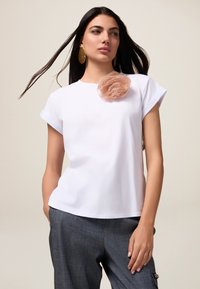Camiseta blanca de manga corta con un acento de flor de tela en suave rosa. Llevada con pantalones grises, que presentan un ajuste relajado y una textura suave.