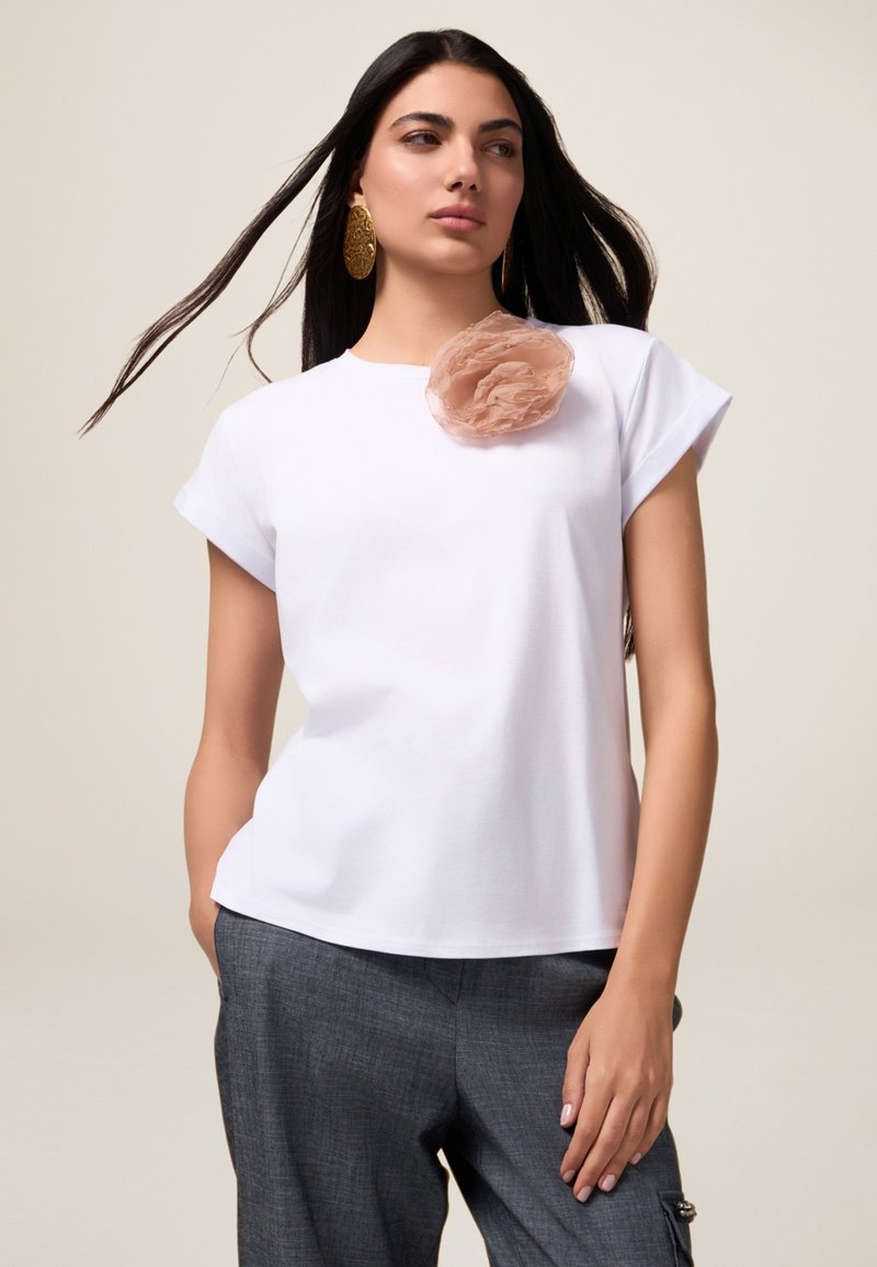 Camiseta blanca de manga corta con un acento de flor de tela en suave rosa. Llevada con pantalones grises, que presentan un ajuste relajado y una textura suave.