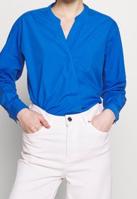 Blaues langärmliges Hemd mit V-Ausschnitt und Wickeldesign, kombiniert mit hellen Hosen. Der Stoff wirkt glatt und leicht glänzend.