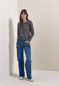 Graues langärmliges Oberteil mit strukturiertem Finish, kombiniert mit blauen weiten Jeans und weißen Sneakers. Der Hintergrund ist hell.