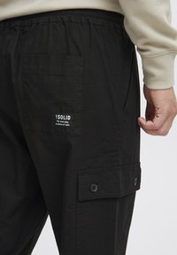 Sorte cargo bukser med tekstureret overflade, der har en elastisk talje, sidelommer og et mærke. Knapdetaljer på cargo-lommen.