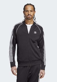 Melna sporta jaka ar rāvējslēdzēju, ar baltām trīs svītru akcentiem uz piedurknēm un Adidas logo uz krūšu daļas. Mīksta tekstūra, pieguloša forma.