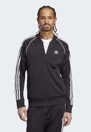 Sort zip-up træningsjakke med hvide tre-stribe accenter på ærmerne og Adidas-logo på brystet. Blød tekstur, tætsiddende design.