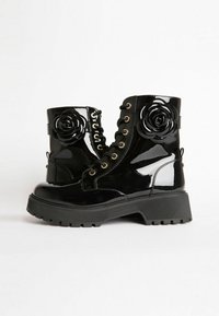 Botas de tobillo de charol negro con adornos decorativos de rosa negra y ojetes en tono dorado, que presentan una suela texturizada gruesa y un diseño de cordones.