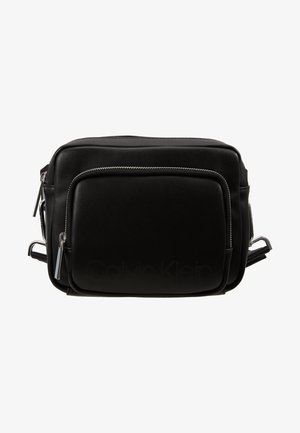 Musta nahkainen Calvin Klein olkalaukku etupaneelin vetoketjulla ja säädettävällä olkahihnalla valkoista taustaa vasten.