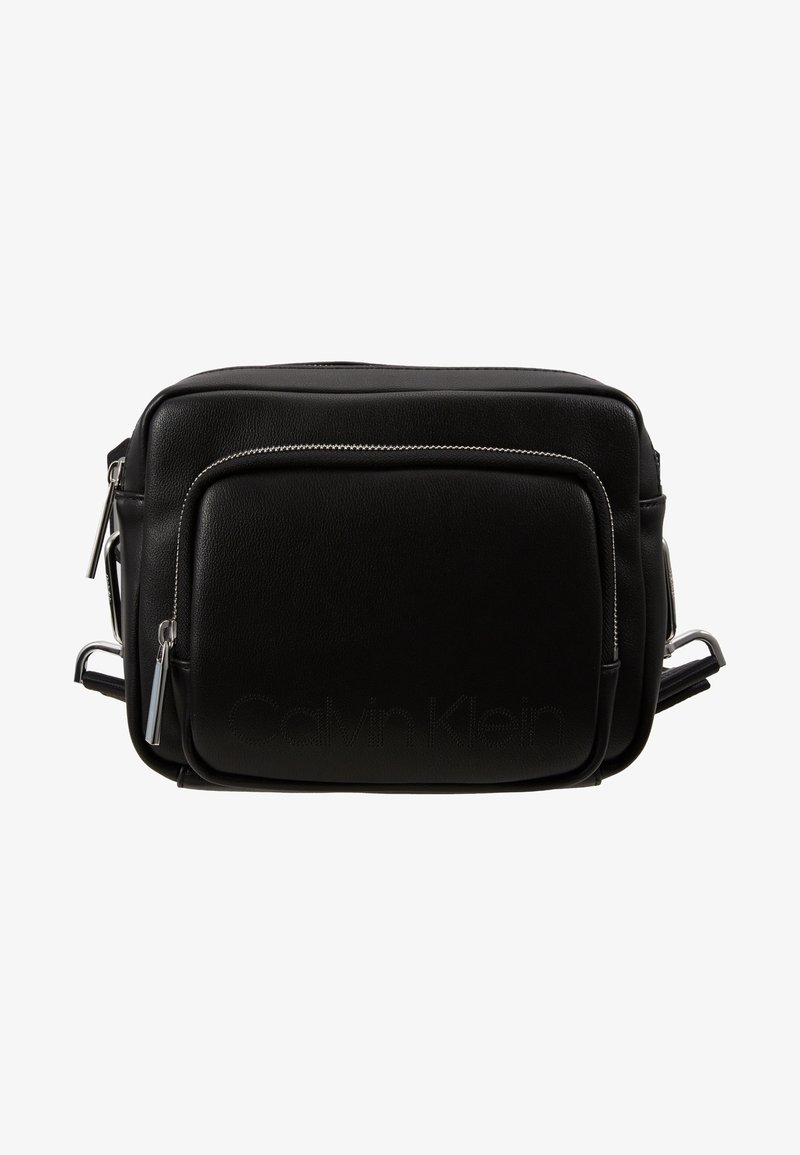 Czarna skórzana torebka crossbody Calvin Klein z przednią kieszenią na zamek i regulowanym paskiem na ramię na białym tle.