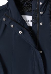 Veste bleu marine foncé avec fermeture éclair et boutons-pression argentés, partiellement ouverte pour montrer la doublure en filet noir et l'étiquette blanche avec le texte de la marque.