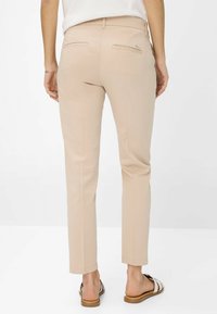 Pantalon beige à coupe slim, fabriqué en tissu lisse. Présente deux poches arrière et un accent logo en métal discret. Porté avec des sandales à enfiler.