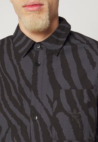 Black and gray shirt with a vertical stripe pattern, button-up design, collar, and a front pocket featuring an embroidered logo.