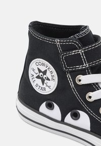 Zapatilla alta de lona negra con ojales, cordones y suela de goma blanca. Presenta un parche del logo de Converse en el lado con un diseño de estrella.