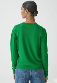 Grøn, strikket sweater med lange ærmer med rynkedetaljer og en rund halsudskæring. Parret med blå denim jeans for kontrast.
