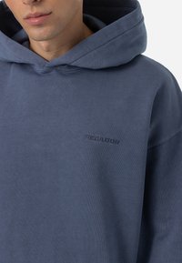 Sudadera con capucha azul oscuro hecha de tela suave y texturizada; cuenta con una capucha grande y el logo bordado "PEGADOR" en el pecho.