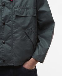 Casaco impermeável verde-escuro com fecho de botões na frente, um bolso no peito e detalhe do logótipo cosido na lateral. Corte casual com mangas longas.