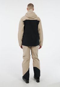 Man in een beige en zwarte skijas en -broek met zwarte skischoenen, staand met zijn rug naar de camera op een witte achtergrond.